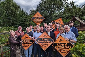 A diverse group of Liberal Democrat campaigners stand outside surrounded by trees and bushes, holding vibrant orange diamond signs featuring the recognisable Liberal Democrat logo and taglines.