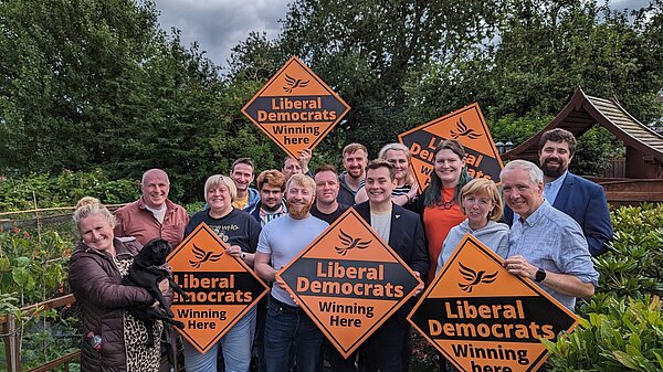 A diverse group of Liberal Democrat campaigners stand outside surrounded by trees and bushes, holding vibrant orange diamond signs featuring the recognisable Liberal Democrat logo and taglines.