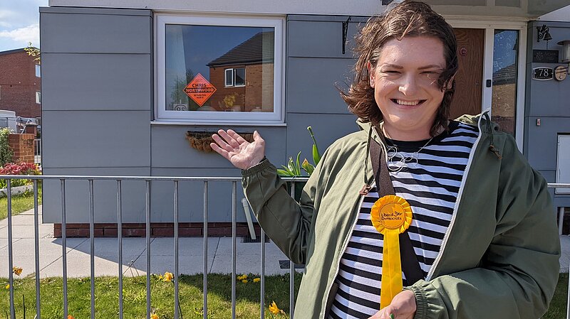 A Lib Dem campaigner stood in front of a house showing a poster in the window