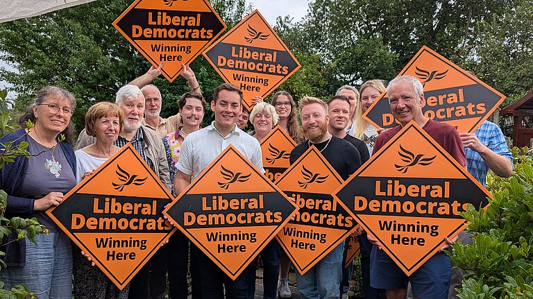 A group of people stand in a garden holding large orange diamond signs saying Liberal Democrats - winning here