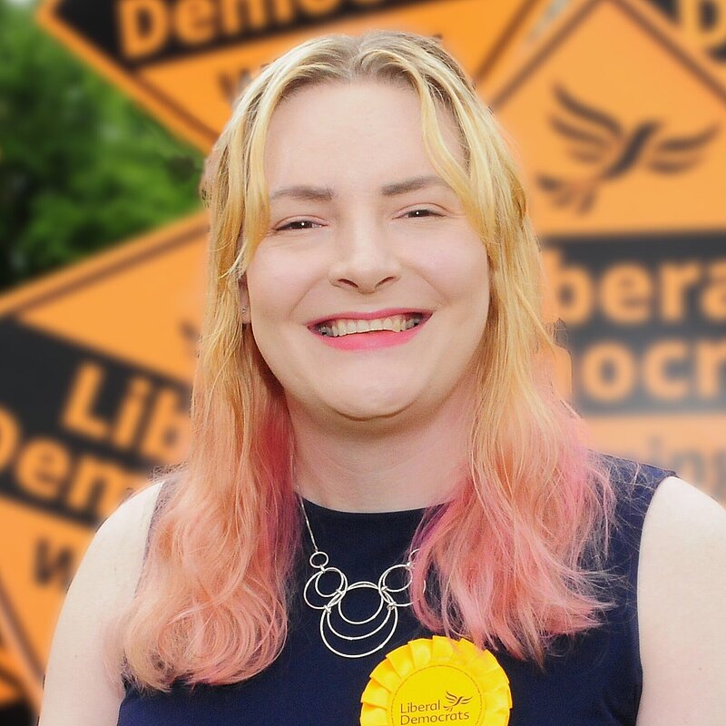 Chris Northwood stands in front a background of people holding large orange diamond signs saying "Liberal Democrats winning here"