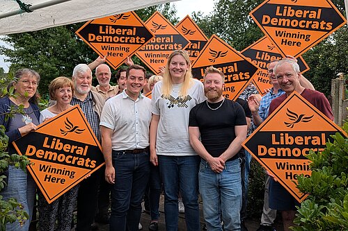 A group of Liberal Democrat campaigners stand outside surrounded by trees and bushes, holding vibrant orange diamond signs featuring the recognisable Liberal Democrat logo and taglines.
